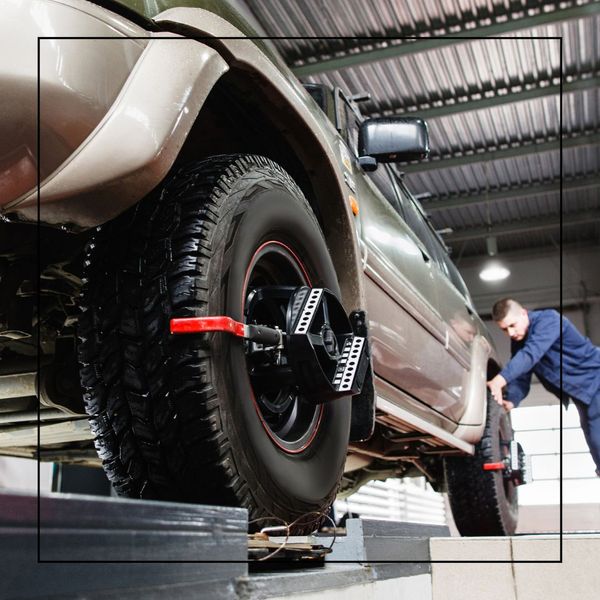 A large, light-colored SUV is raised on a lift for wheel alignment, with a sensor attached to its front tire, and a mechanic leaning over the back wheel in the background. A large, light-colored SUV is raised on a lift for wheel alignment, with a sensor attached to its front tire, and a mechanic leaning over the back wheel in the background.