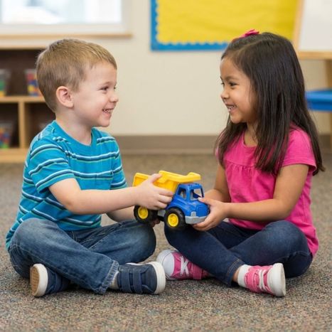 Two smiling pre-k children sharing a toy truck on a carpet.