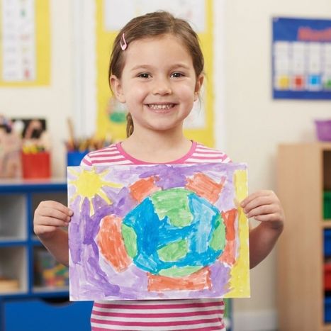 A smiling pre-k girl proudly holding up her painting in a classroom.