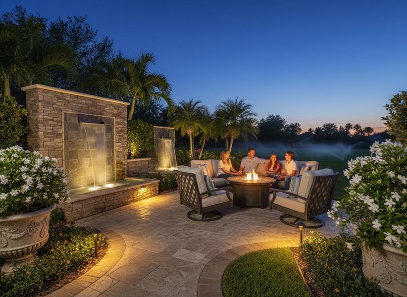 A family of four sits around a fire pit on a stone patio with decorative waterfalls and lush landscaping at twilight. Family enjoying outdoor patio with fire pit and water feature at dusk