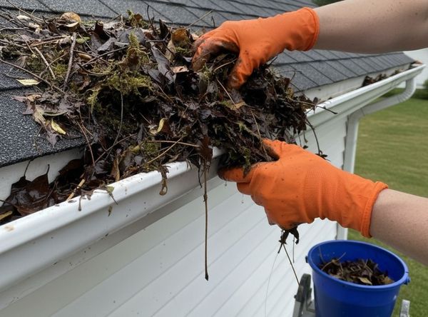 A pair of hands in protective gloves removes a large handful of wet leaves and debris from a clogged residential gutter.