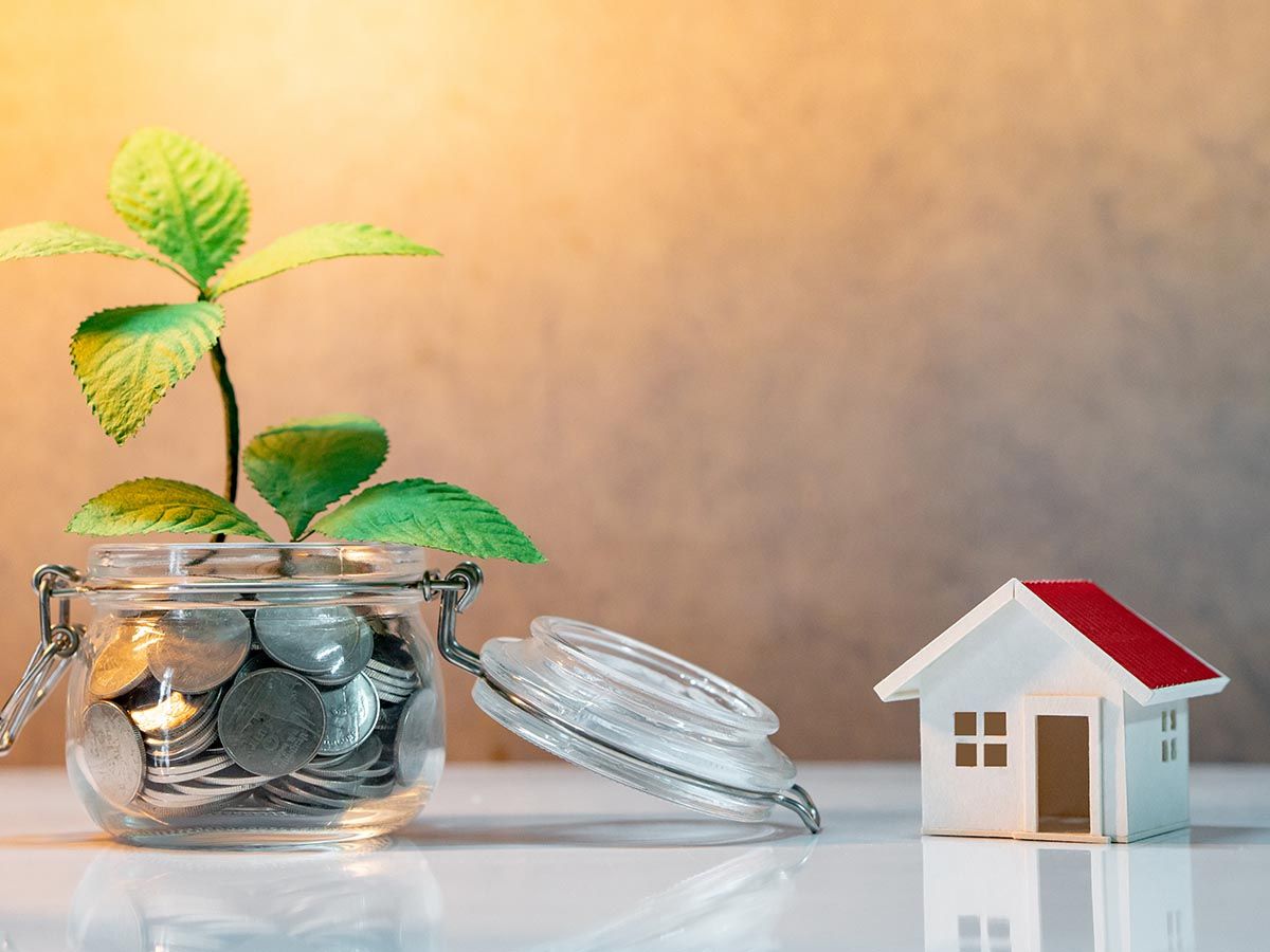 Saving money for future concept. Reflection of green plant growing out of coins in glass jar and house model on the table.