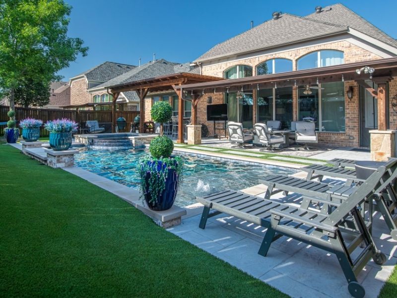 A rectangular inground swimming pool with vibrant blue water, stone coping, and several grey lounge chairs positioned on a light-colored patio.