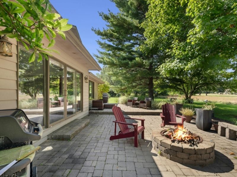 A paved backyard patio featuring a circular stone fire pit with two red Adirondack chairs.