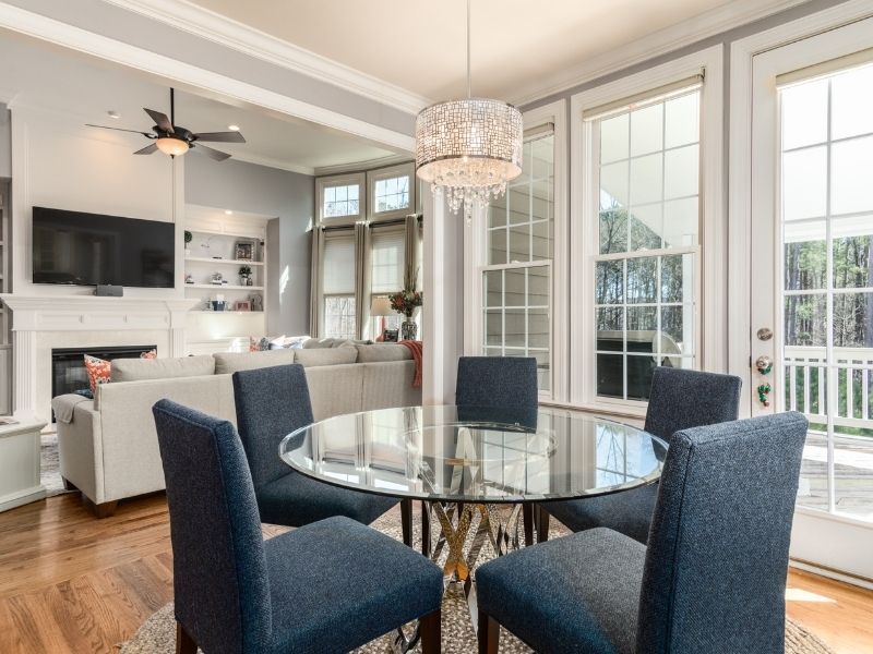 A round glass-top dining table with a metallic base surrounded by four navy blue upholstered chairs under a crystal chandelier.