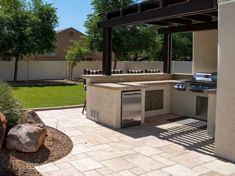 An outdoor kitchen area featuring a stainless steel grill, a mini-refrigerator, and a sink built into a stone-clad counter under a dark pergola.