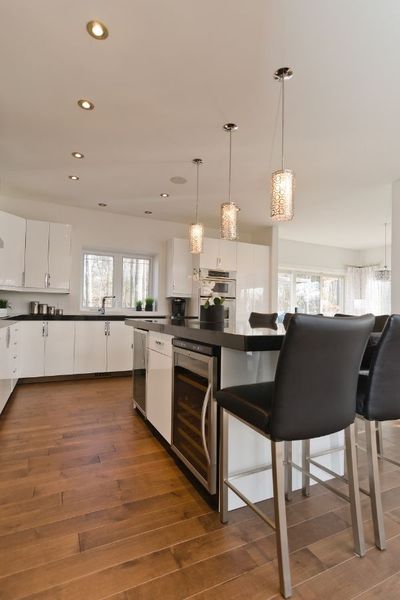 A contemporary kitchen island with a dark countertop, a built-in wine cooler, and black leather bar stools situated under three decorative hanging pendant lights.