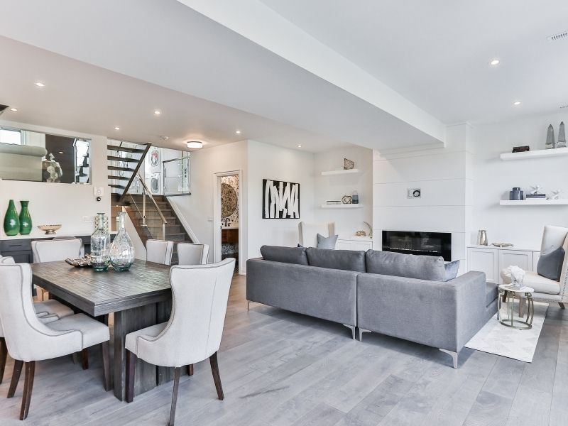 A modern open-concept living and dining area featuring a dark wood dining table with high-back upholstered chairs and a large grey sectional sofa facing a minimalist white fireplace.