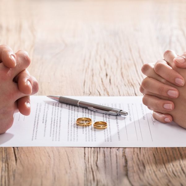 Two people with their hands crossed over a piece of paper, a pen, and two wedding rings.