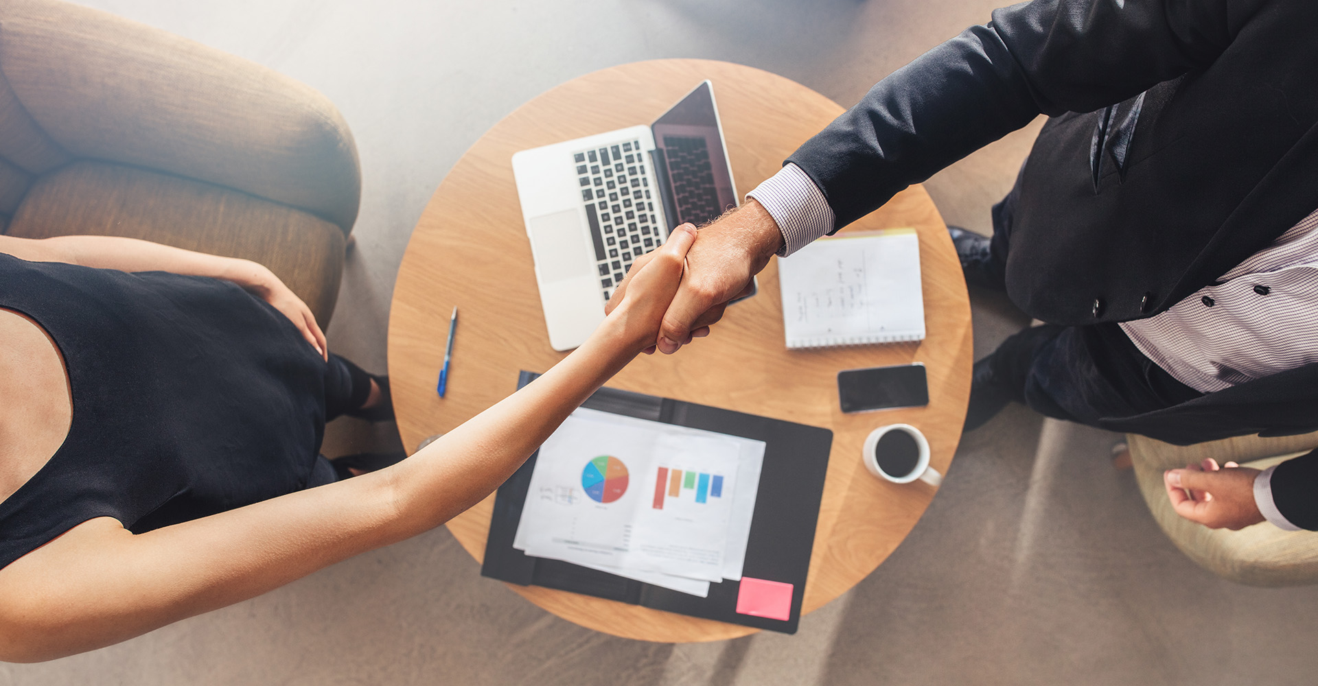 Two people shaking hands over a table with paperwork and a computer on it