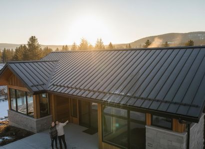 Aerial view of a modern home with a dark standing seam metal roof and wooden accents. A couple points at the roof on the patio, with snowy mountains in the background under a bright sun. Modern House with Standing Seam Metal Roof and Mountain View