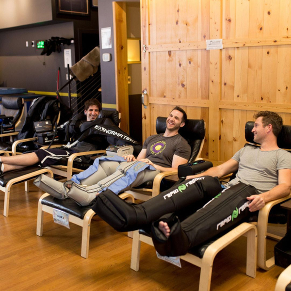 Three athletes sit in a recovery lounge using professional pneumatic leg compression systems.