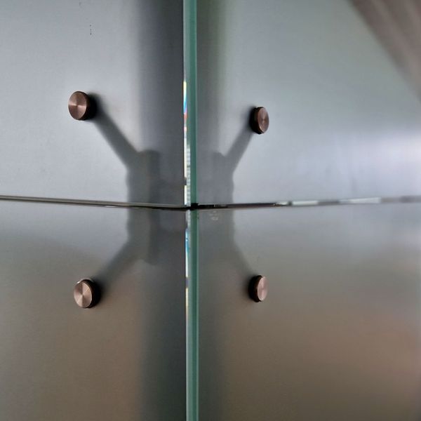 A detailed close-up of four brushed bronze standoff fittings securing the corner of a frosted glass partition wall.