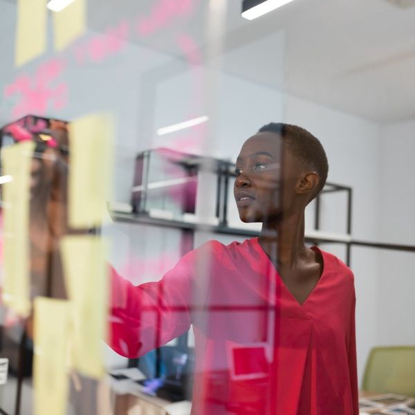A woman in a red top placing yellow sticky notes on a clear glass partition, brainstorming or organizing ideas in an office setting.