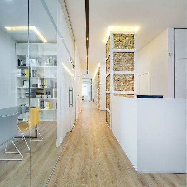 Modern office hallway with light wooden flooring, glass partitions leading to an office space on the left, and a white reception desk on the right with a stone-clad pillar.