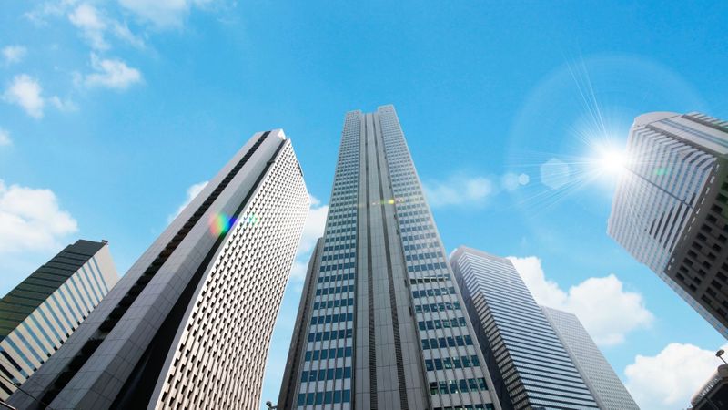 Low-angle view of multiple modern high-rise buildings with glass facades against a bright blue sky with scattered clouds, and a sun flare. Low-angle view of multiple modern high-rise buildings with glass facades against a bright blue sky with scattered clouds, and a sun flare.