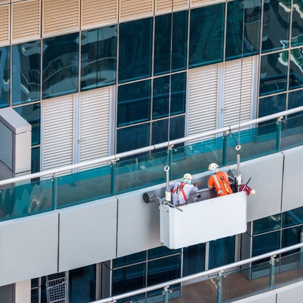 Two construction workers in safety gear and hard hats working from a suspended platform, performing maintenance on the exterior windows of a commercial building.