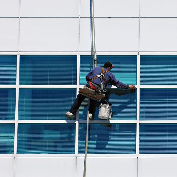 Window washer suspended by ropes, cleaning the exterior windows of a modern high-rise building with blue tinted glass.