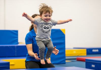 A happy toddler in a grey t-shirt with a raccoon graphic leaps through the air, arms outstretched, assisted by an adult in a blue shirt, in a gymnastics setting with blue and yellow mats. The child is smiling, enjoying the sensation of flight and movement. Toddler Jumping with Assistance