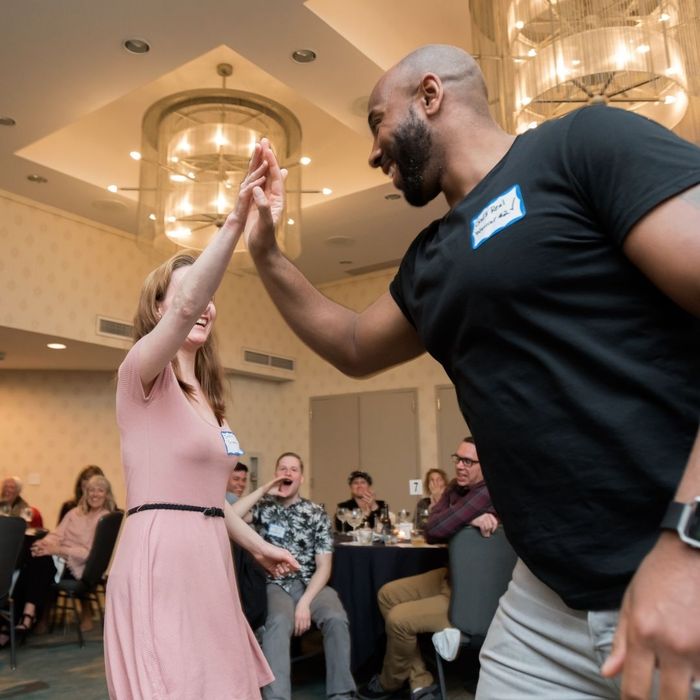 A man and a woman in the foreground happily high-five at a dinner show, celebrating a moment while guests seated at tables watch and smile in the background. A man and a woman in the foreground happily high-five at a dinner show, celebrating a moment while guests seated at tables watch and smile in the background.