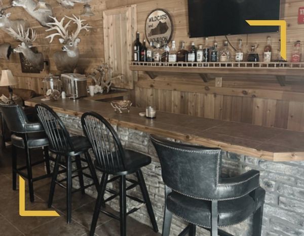 An indoor bar area at the hunting lodge with a stone-faced counter, leather barstools, a selection of spirits, and decorative whitetail mounts.