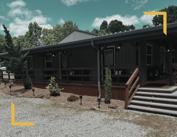 The exterior of the Kentucky hunting lodge in Olaton, featuring a large covered wooden porch with steps leading to a gravel driveway under a bright blue sky.