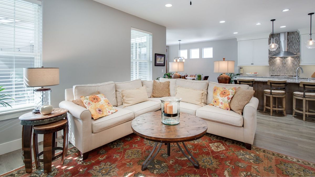 A bright, open-concept living room featuring a cream sectional sofa, a rustic round wood coffee table, and an ornate red area rug, transitioning into a modern kitchen in the background.