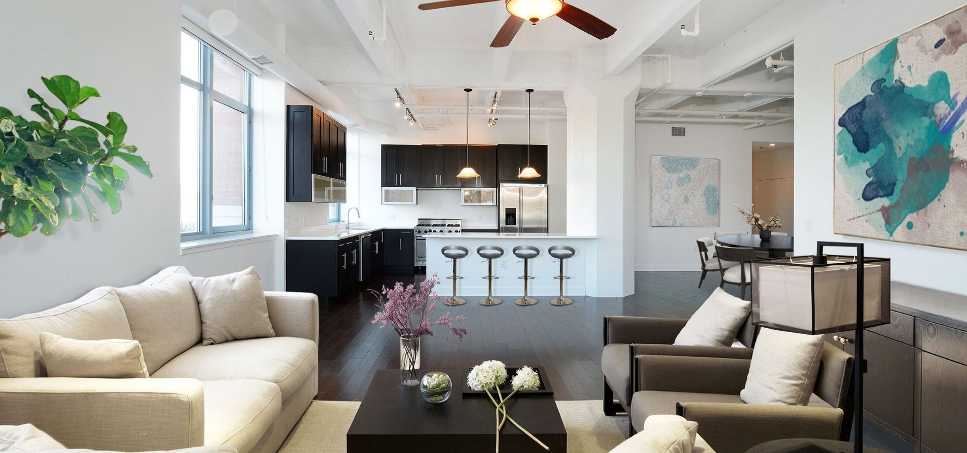 Industrial-style loft renovation featuring a dark modern kitchen, exposed white ceiling pipes, and a bright, open living room with dark wood flooring.