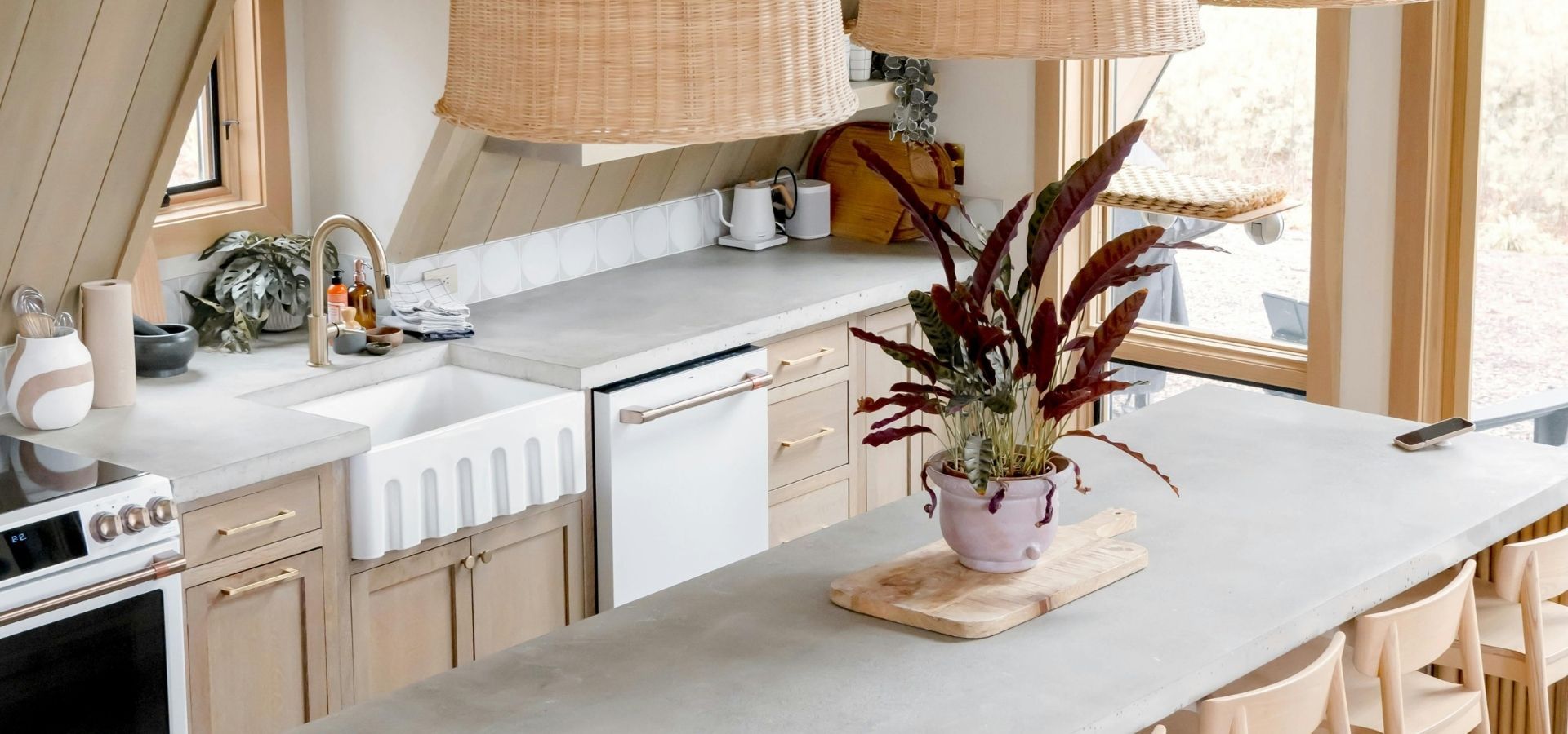 Close-up of a modern rustic kitchen remodel featuring smooth concrete countertops, a white fluted farmhouse sink, and light wood cabinetry with gold hardware.