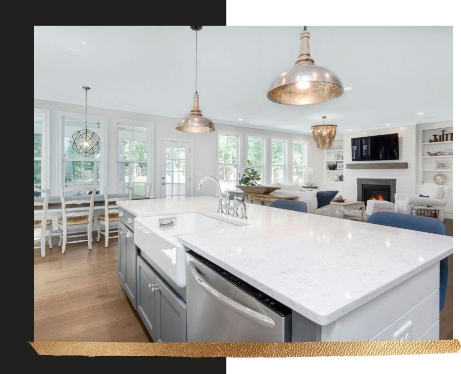 Large open-concept kitchen featuring a white marble island with a farmhouse sink, grey cabinetry, and industrial pendant lighting overlooking a bright living room.