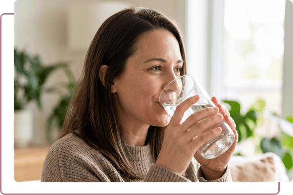 A smiling woman indoors holding a clear glass and drinking water.