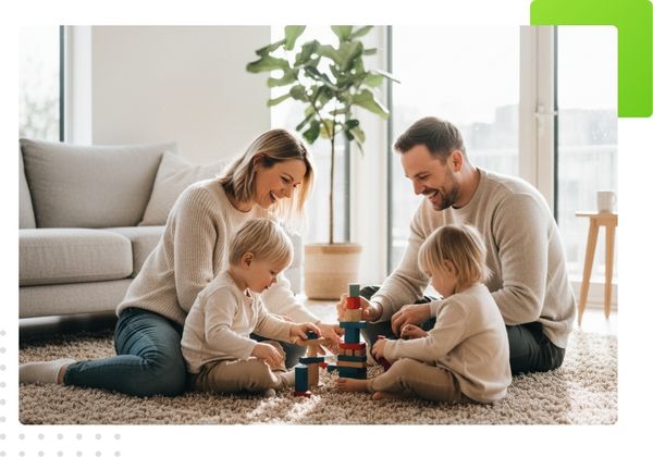 A smiling family of four sitting on a light-colored rug, building a tower with colorful wooden blocks.