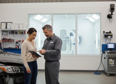 A service advisor points to a tablet while talking to a customer next to a damaged car, with a painter in a booth behind them. Mechanic and Customer Reviewing Car Repair Details