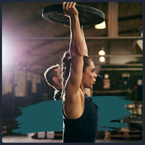 Woman holding a weight plate above her head in a brightly lit gym.