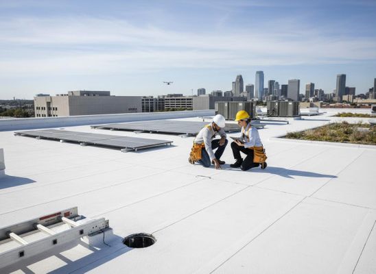 Two engineers in hard hats inspecting a commercial rooftop with solar panels and a city skyline. A drone flies overhead. Rooftop Inspection with Engineers and Solar Panels