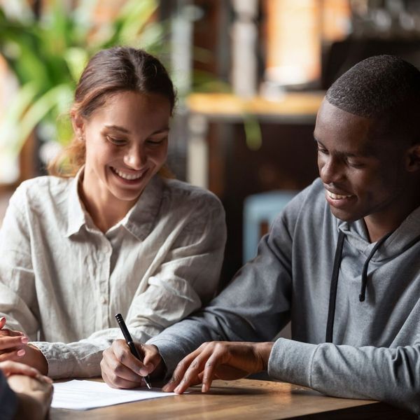 A young couple, one smiling woman and one smiling man, happily signing a document together at a table.