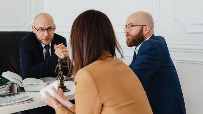 A lawyer speaking to two clients (a man and a woman) in an office setting, with a small Lady Justice statue on the desk.