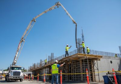 Construction workers in hard hats and safety vests oversee a concrete pour on a construction site, with a large concrete pump truck extending its boom to deliver the material to a reinforced structure under a clear blue sky. Safety cones and caution tape mark the perimeter of the work zone. Concrete Pouring at Construction Site