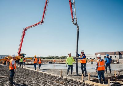 A group of construction workers wearing safety vests and hard hats are working on a construction site. A large concrete pump truck with a red boom is pouring concrete onto a rebar grid. Some workers are guiding the concrete hose while others observe the process. Construction workers pouring concrete on a construction site.