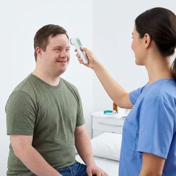 A female nurse uses a forehead thermometer to check the temperature of an adult male with Down syndrome during a routine health monitoring check.