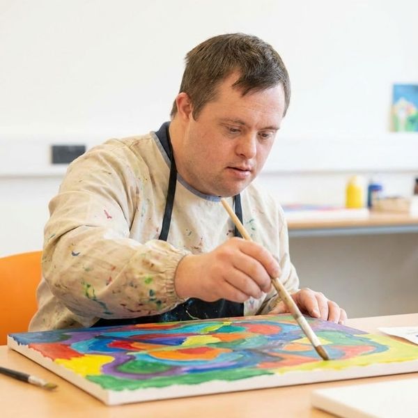 An adult man with Down syndrome is intently focused on painting a colorful canvas during an art activity at an adult day care center.
