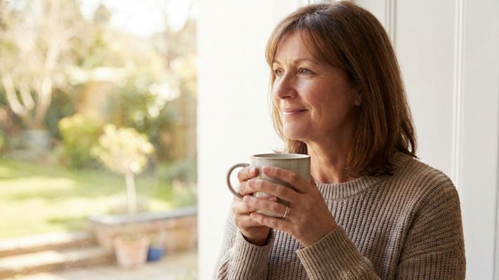 Woman smiling and holding a mug, looking out a window in a moment of peace.