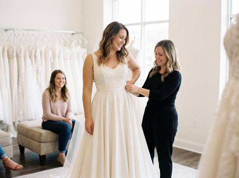 A happy bride-to-be smiles while a consultant adjusts her white wedding dress in a bright bridal shop, with a friend watching nearby. Bride-to-be tries on wedding dress with consultant