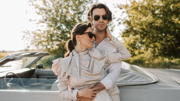A stylish couple wearing sunglasses posing together in front of a classic white convertible car on a bright, sunny day with trees in the background. A stylish couple wearing sunglasses posing together in front of a classic white convertible car on a bright, sunny day with trees in the background.