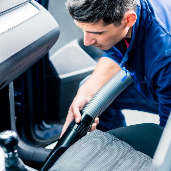 A professional detailer in a blue uniform meticulously using a vacuum attachment to deep clean the interior fabric seats of a vehicle.