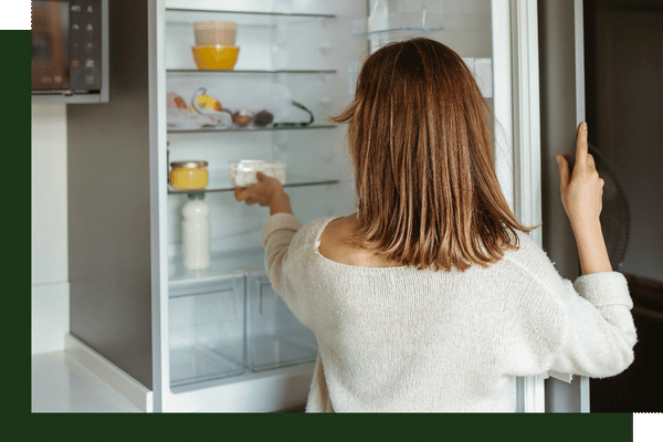 A woman with her back turned looking inside a fully stocked, well-lit white refrigerator with the door wide open.