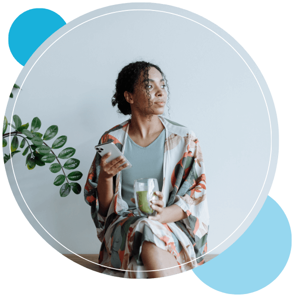 A person wearing a patterned robe sits while holding a smartphone in one hand and a glass of green juice in the other.
