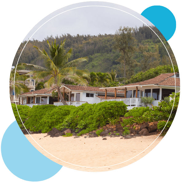Beachfront houses with lanais sit on a sandy shore behind a line of thick green shrubbery.