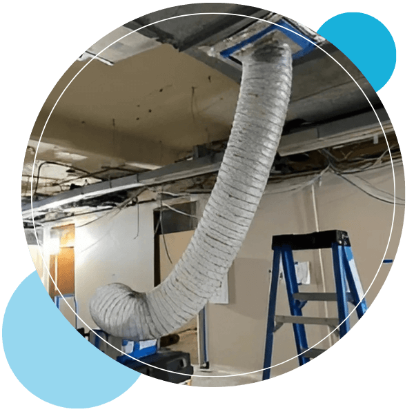 A large, flexible ventilation duct cleaner hangs down from an unfinished ceiling with exposed framework.