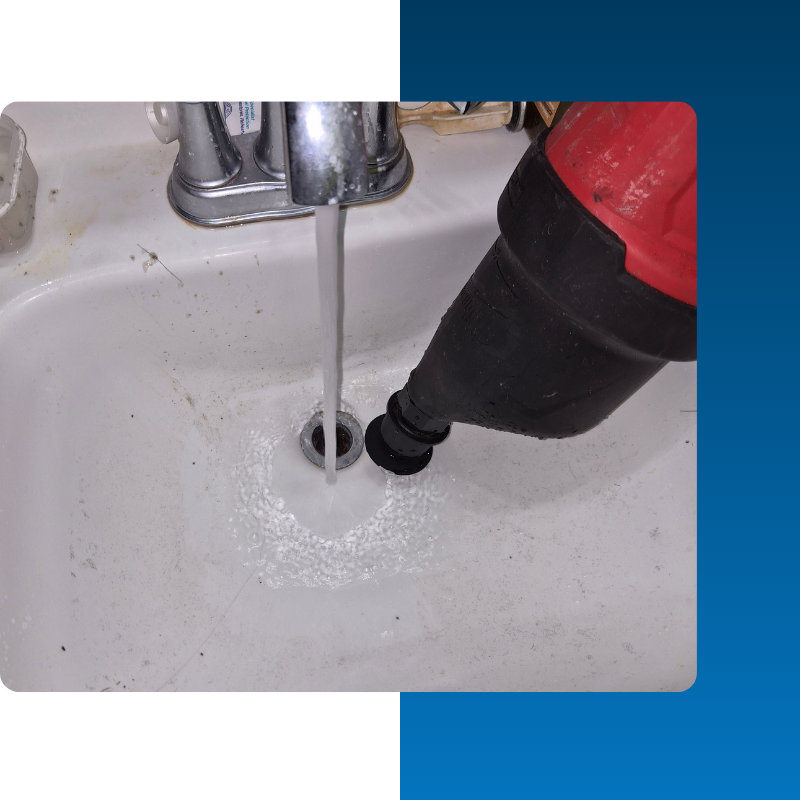 A handheld power drain cleaner (drain auger) is positioned over a white bathroom sink drain while water runs from the faucet. The red and black tool is used to clear small clogs in sink traps and secondary drain lines.